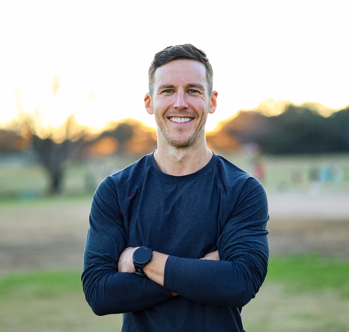 Corey standing outdoors with arms crossed, wearing a dark long-sleeve shirt and a smartwatch, with a sunset-lit park in the background.